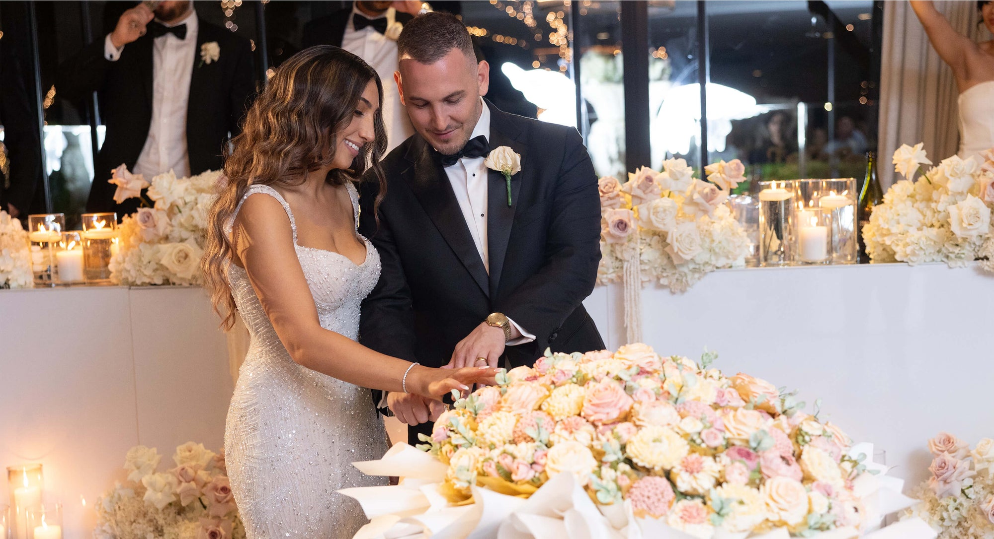 bride and groom cutting flower wedding cake with florals and candles in background