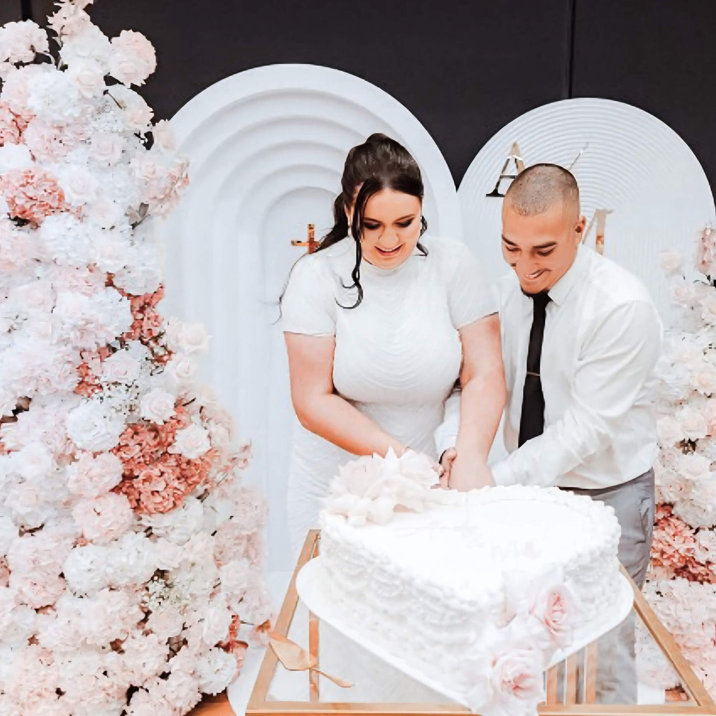 bride and groom cutting wedding cake in front of white backdrops and pink and white flower tower