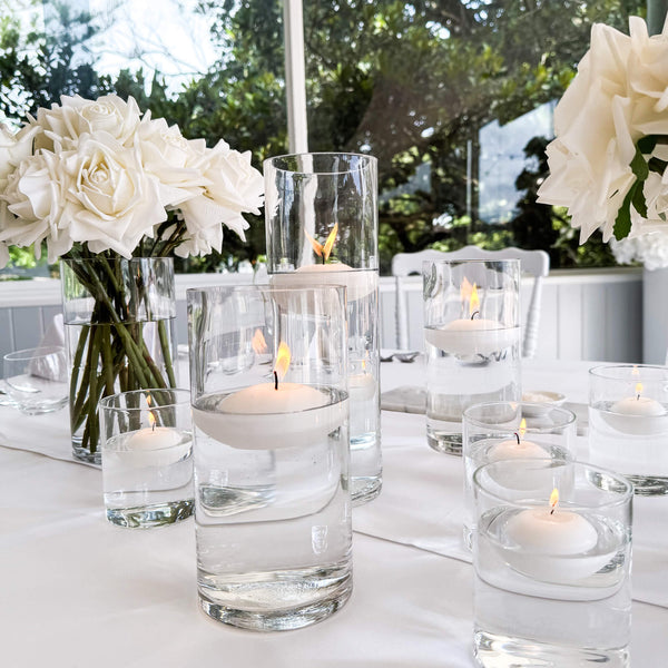 Table setting with floating water candles in glass holders and white roses on a white tablecloth.