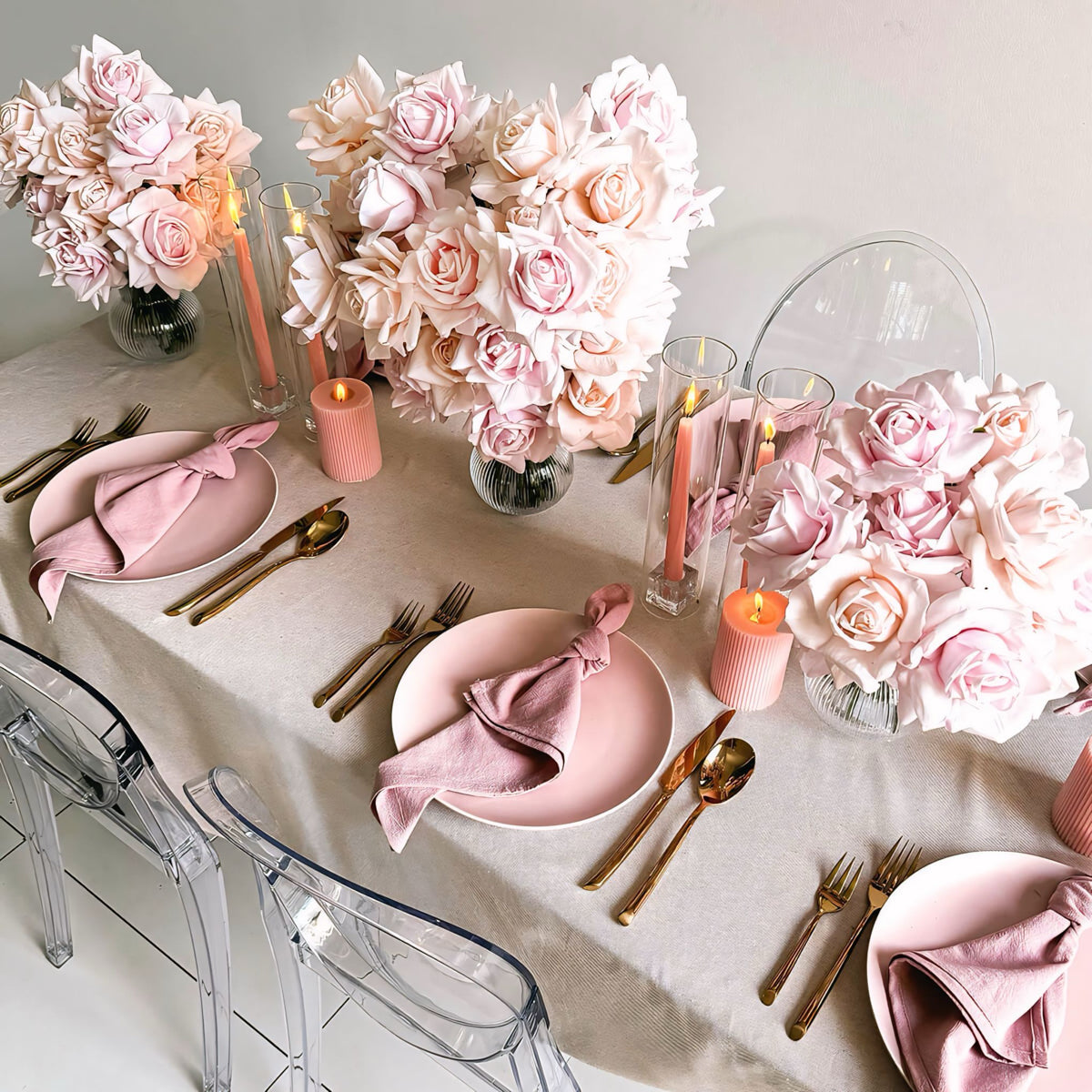 three pink rose flower arrangements on beige tablecloth with pink candles and rose gold cutlery in between
