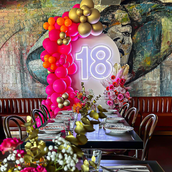 white 18 neon sign hanging on arch backdrop with pink and gold balloon arch and flowers