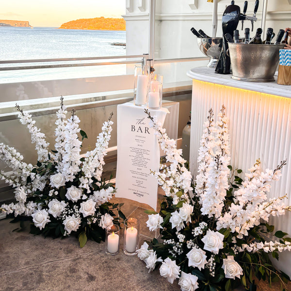 Decorative bar sign setup with flowers, candles, and a 'The Bar' menu sign by a waterfront.