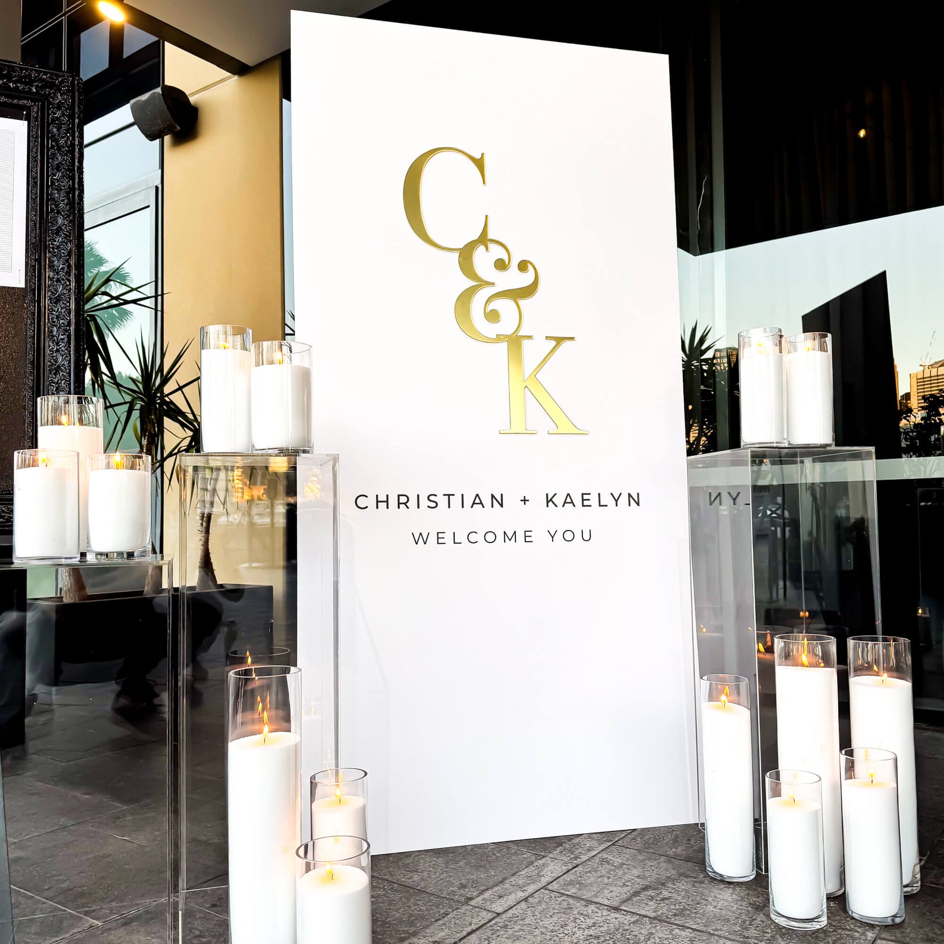 white rectangle welcome sign with couples C & K initials, in between white sand candles placed on clear plinths