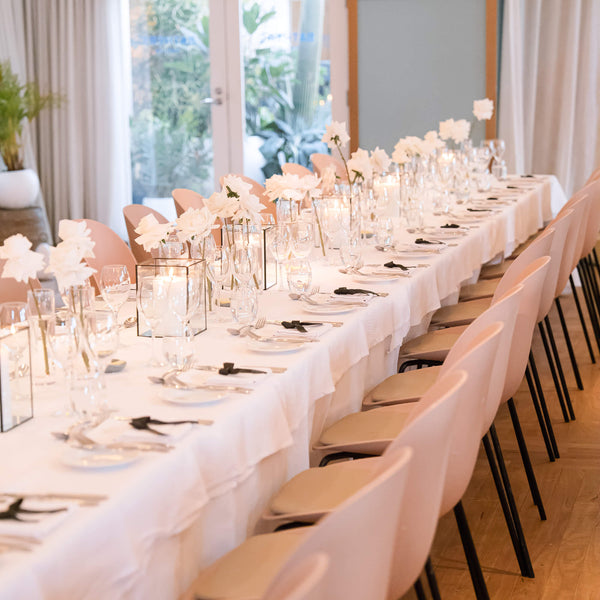 Long table set for a wedding event with white tablecloths, white rose floral centerpieces, black candle boxes and chairs.