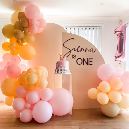 beige arch backdrop setup with pink and peach balloons at child's first birthday