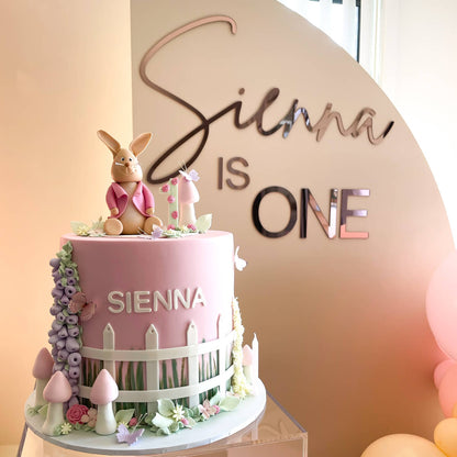 pink flopsy birthday cake with rose gold acrylic lettering attached to beige backdrop in background