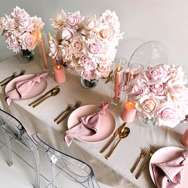 three pink rose flower arrangements on beige tablecloth with pink candles and rose gold cutlery in between