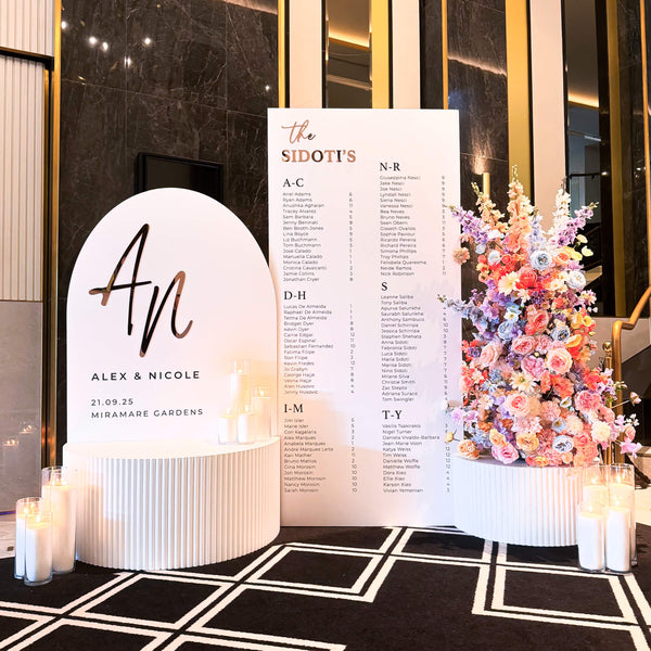 Decorative wedding sign setup with a floral arrangement, sand candles, seating chart, and personalised welcome sign in an elegant interior setting.